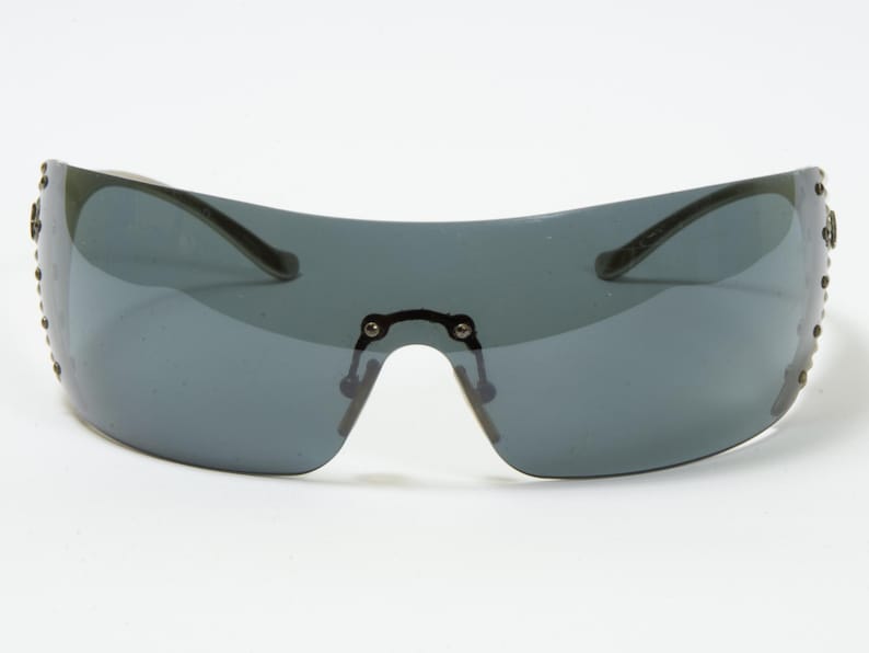 May include: A pair of shield sunglasses with a dark gray lens and a curved, wraparound design. The frame is a dark color, and small decorative studs line the outer edges. The sunglasses have a bridge and arms.