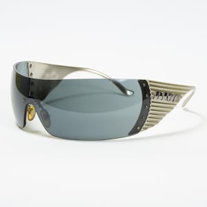 May include: Silver-toned sunglasses with a wraparound design and dark gray lenses. The sunglasses feature a unique, futuristic style with a ridged detail on the temples and a curved frame. The lenses are a solid, dark gray color.