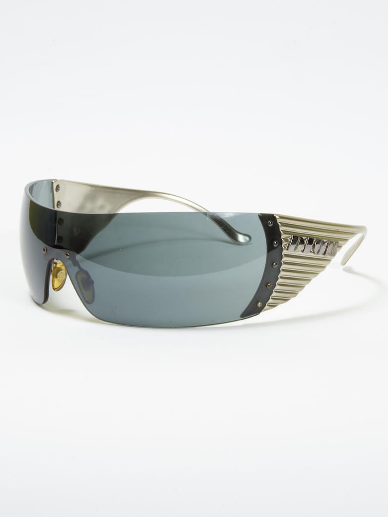 May include: Silver-toned sunglasses with a wraparound design and dark gray lenses. The sunglasses feature a unique, futuristic style with a ridged detail on the temples and a curved frame. The lenses are a solid, dark gray color.