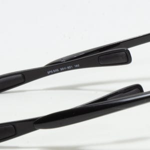 May include: Close-up of black eyeglasses arms. The arms have a rectangular pad at the end for comfort. The text "SPS 50G 8AV-501 140" is printed on the arm. The glasses are set against a white background.