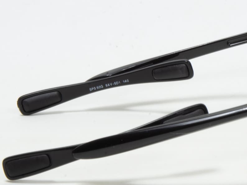 May include: Close-up of black eyeglasses arms. The arms have a rectangular pad at the end for comfort. The text "SPS 50G 8AV-501 140" is printed on the arm. The glasses are set against a white background.