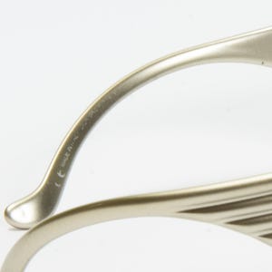 May include: Close-up of a pair of eyeglasses with a metallic, light gold frame. The temples curve gracefully, and the frame has a ridged design. The text "MADE IN ITALY" is visible on the temple.