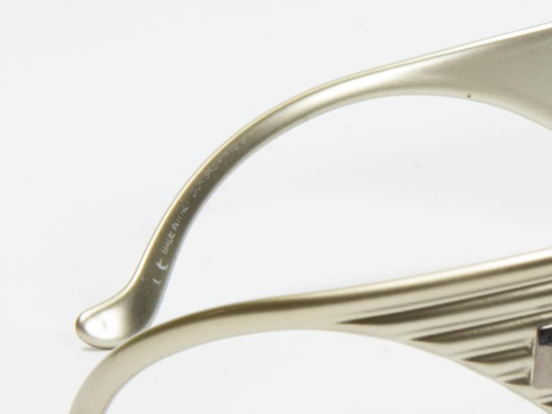 May include: Close-up of a pair of eyeglasses with a metallic, light gold frame. The temples curve gracefully, and the frame has a ridged design. The text "MADE IN ITALY" is visible on the temple.