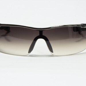 May include: Black sports sunglasses with a gradient lens. The sunglasses have a curved, wraparound design with a black frame and a small red accent on the temple. The lenses transition from dark at the top to light at the bottom.