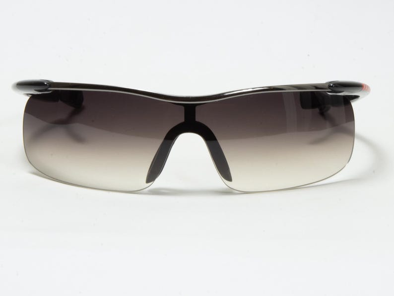 May include: Black sports sunglasses with a gradient lens. The sunglasses have a curved, wraparound design with a black frame and a small red accent on the temple. The lenses transition from dark at the top to light at the bottom.