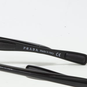 May include: Close-up of black Prada sunglasses. The temple arm features the brand name and "Made in Italy" text. The sunglasses have a sleek, modern design with a red accent on the arm.