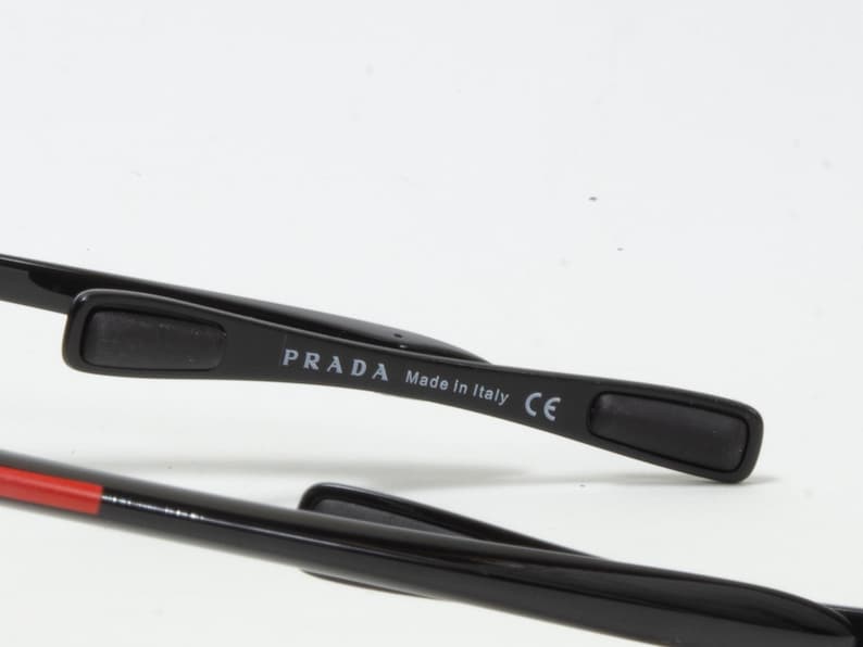 May include: Close-up of black Prada sunglasses. The temple arm features the brand name and "Made in Italy" text. The sunglasses have a sleek, modern design with a red accent on the arm.