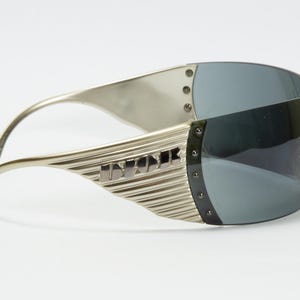 May include: Silver-toned sunglasses with a wraparound design. The lenses are a dark gray, and the frames feature a ridged texture with the word "DIOR" in silver lettering. The sunglasses have a futuristic aesthetic.