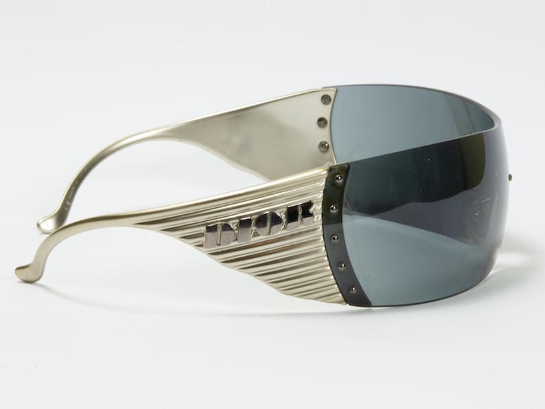 May include: Silver-toned sunglasses with a wraparound design. The lenses are a dark gray, and the frames feature a ridged texture with the word "DIOR" in silver lettering. The sunglasses have a futuristic aesthetic.