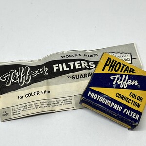 May include: A yellow and blue box with the text "Tiffen Photar Color Correction Photographic Filter" on it. The box is sitting on top of a folded piece of paper with the text "Tiffen" and "World's Finest Filters" on it.