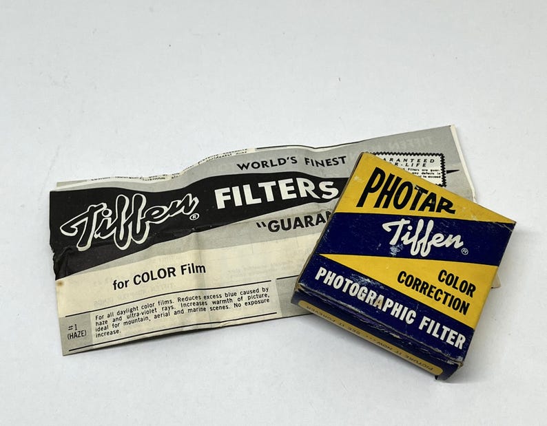 May include: A yellow and blue box with the text "Tiffen Photar Color Correction Photographic Filter" on it. The box is sitting on top of a folded piece of paper with the text "Tiffen" and "World's Finest Filters" on it.