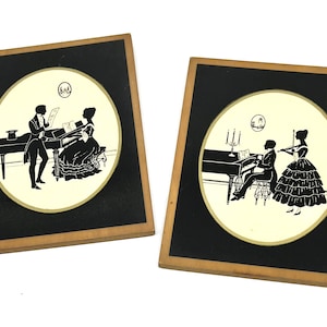 May include: Two rectangular wall plaques with light wood frames, each featuring a black and cream circular silhouette illustration. One depicts a person playing piano while another stands, and the other shows a person playing piano with a violinist.