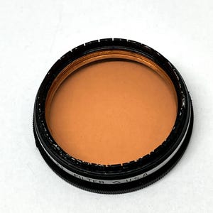 May include: A round, black camera filter with a brown glass lens. The filter has the text "FILTER" and "USA" printed on the side.