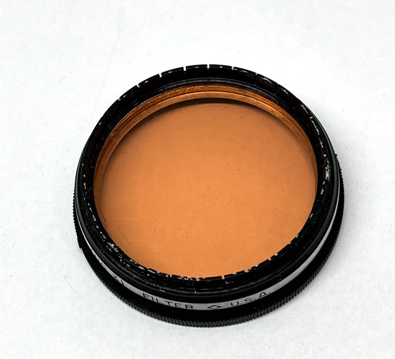 May include: A round, black camera filter with a brown glass lens. The filter has the text "FILTER" and "USA" printed on the side.