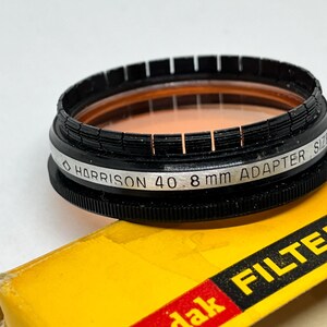 May include: A black and silver camera filter with the text "HARRISON 40 8 mm ADAPTER SIZE" printed on the side. The filter is sitting on a yellow box with the text "Kodak FILTER" printed in red.