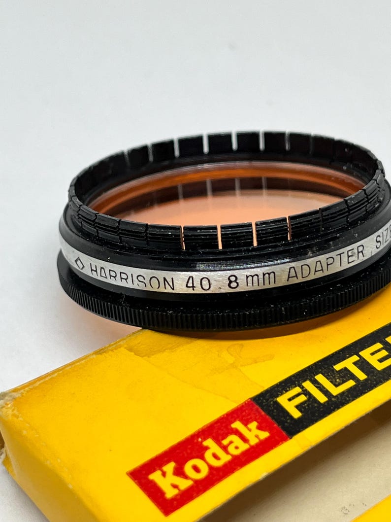 May include: A black and silver camera filter with the text "HARRISON 40 8 mm ADAPTER SIZE" printed on the side. The filter is sitting on a yellow box with the text "Kodak FILTER" printed in red.