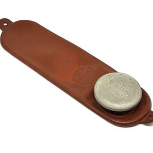 May include: A brown leather case with a circular metal lid. The lid is silver with the text "Wurless" and other text. The case has a loop at one end and a circular emblem.