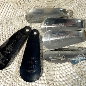 May include: A collection of vintage shoe horns in various shapes and sizes. Some are black, others are silver-toned metal. Several have text, including "FREEMAN SHOE FOR MAN" and "FRIEDMAN SHELBY ALL LEATHER SHOES".