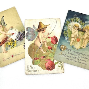 May include: Three vintage Valentine's Day postcards. One features a cherub with a scroll, another shows a cherub fishing with a heart-shaped lure, and the third depicts two cherubs with a heart. Each card has text.