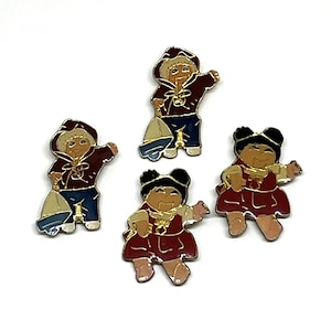 May include: Four enamel pins featuring children in various outfits. Two pins depict children in hooded jackets and holding sailboats. The other two pins show children in dresses. All pins have gold-colored trim.