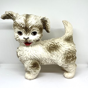 May include: A vintage-style puppy figurine in a standing pose. The dog has a white and tan color scheme with textured fur, large brown eyes, and a red mouth. The figurine is made of a hard material and has a glossy finish.