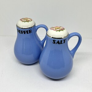 May include: A pair of blue ceramic salt and pepper shakers. Each shaker has a handle and a floral design on the lid. The word "PEPPER" is printed on one shaker, and "SALT" is printed on the other.
