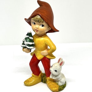 May include: A ceramic figurine of a young elf-like figure wearing a brown hat, yellow tunic, and red pants, holding a small green Christmas tree. A white rabbit sits at the elf's feet. The figurine has a vintage, whimsical style.
