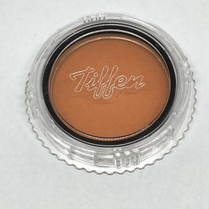 May include: A round, orange-coloured filter in a clear plastic container with the word "Tiffen" printed on the filter.