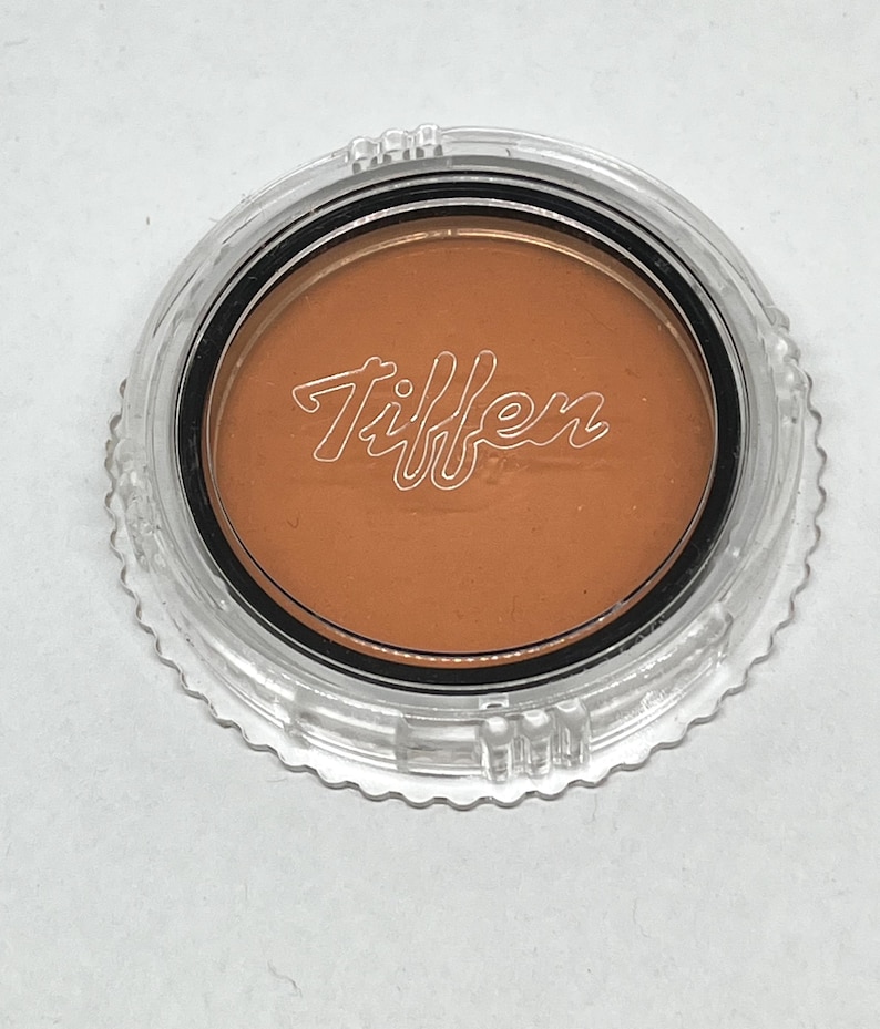 May include: A round, orange-coloured filter in a clear plastic container with the word "Tiffen" printed on the filter.