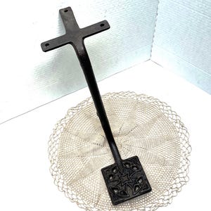 May include: A dark metal decorative cross-shaped object with a long handle and a square base featuring a star and floral design. The object is placed on a cream-colored, crocheted doily.