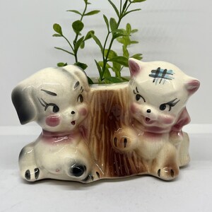May include: A vintage ceramic planter featuring a dog and cat design. The dog has a black ear and the cat has a blue cross on its head. The planter is tan, brown, and cream-colored, with pink accents. Green plants are inside.