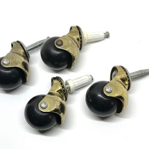 May include: Four antique brass and black furniture casters. Each caster has a black ball wheel, a brass-colored metal housing, and a silver-colored stem. The casters are designed for furniture and other applications.