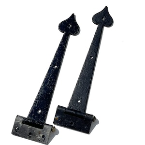 May include: Two black, wrought iron door hinges. Each hinge features a heart-shaped finial and a rectangular base with screw holes. The hinges are designed for durability and a classic aesthetic.
