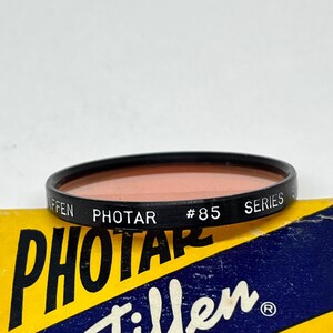 May include: A Tiffen Photar #85 Series filter in a black plastic ring with a peach-coloured glass lens. The filter is sitting on top of a yellow and blue box with the text "Photar Tiffen" printed on it.