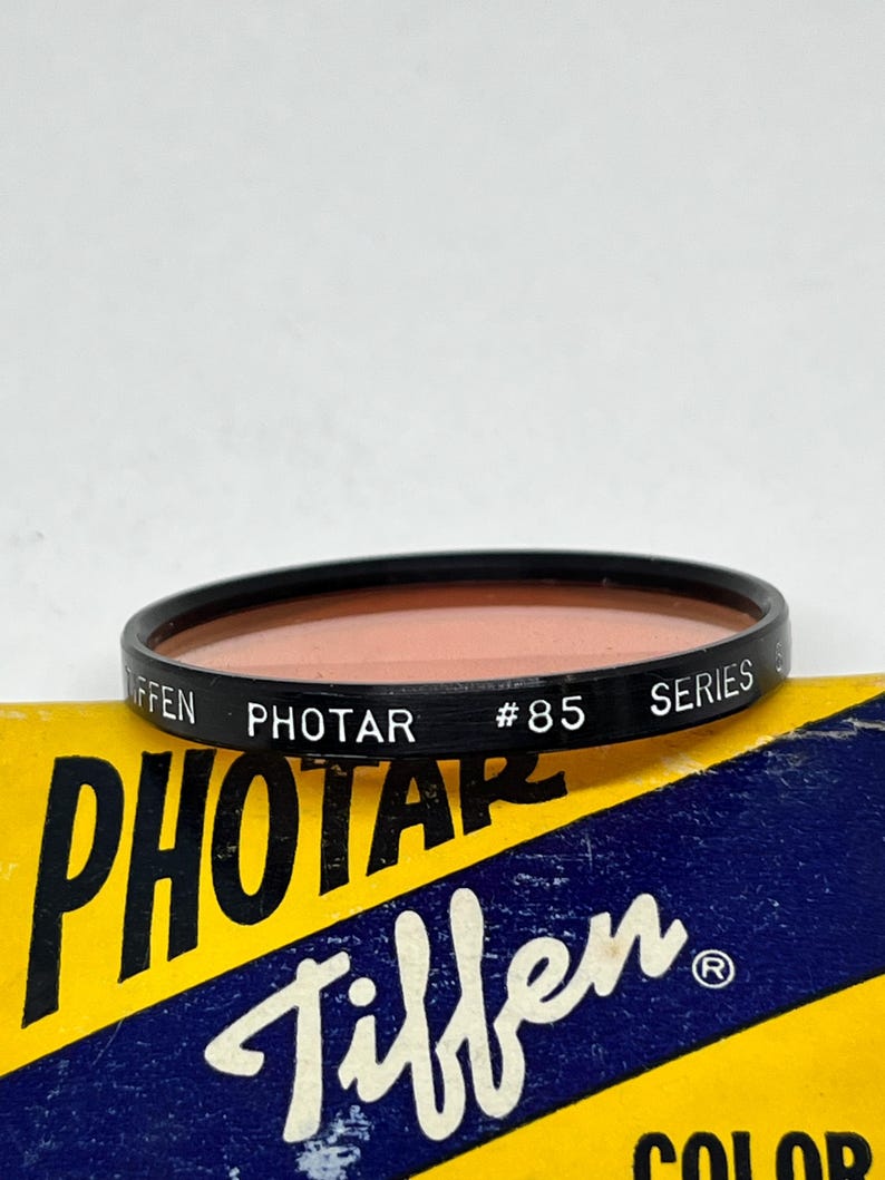 May include: A Tiffen Photar #85 Series filter in a black plastic ring with a peach-coloured glass lens. The filter is sitting on top of a yellow and blue box with the text "Photar Tiffen" printed on it.