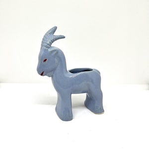 May include: A light blue ceramic goat figurine with red eyes and a red mouth. The goat has large curved horns and a hollowed-out back, suggesting it is a planter or decorative item. The figurine is on a white background.