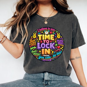 Può includere: T-shirt grigio scuro con una grafica colorata che recita "Time to Lock In". Il design include le frasi "Rock the test", "Don't stress" e "You can do this?" in vari font e colori. La maglietta ha uno scollo tondo.