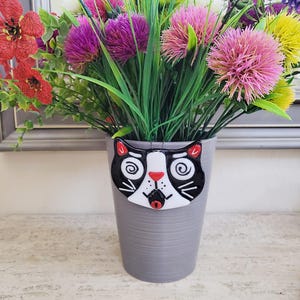 May include: A gray ceramic flower pot with a whimsical cat face design. The cat face is black and white with red accents, and the pot is filled with colorful artificial flowers. The pot is a decorative item.