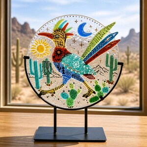May include: A circular glass art piece featuring a colorful roadrunner bird, a yellow sun, a blue crescent moon, and green cacti. The design includes intricate patterns on the bird's body and is supported by a black metal stand on a flat base.