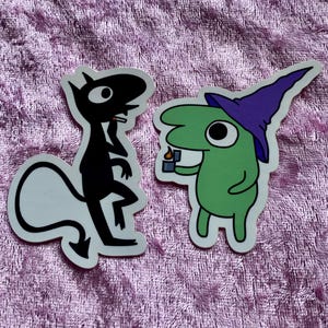 May include: Two die-cut stickers featuring cartoon characters. One is a black devil-like figure with a white tail, and the other is a green creature wearing a purple witch hat and holding a small candle. The stickers are on a pink, textured surface.