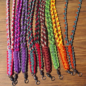 May include: A selection of brightly coloured braided lanyards. Each lanyard displays a unique colour combination, including red, purple, green, and orange. The lanyards are made of a durable material and have a metal clasp at the end.