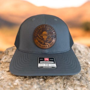 May include: A dark gray and black mesh trucker hat with a brown leather patch that says "Badlands Customs Established in 2023". The patch features a skull with horns and a sunset behind it.