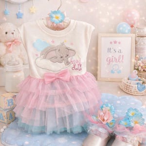 May include: A baby girl's outfit with a white top featuring an elephant design and a pink tutu skirt. The outfit is displayed with matching shoes, a welcome baby sign, and a framed "It's a girl!" print. The color scheme is soft pink and blue.