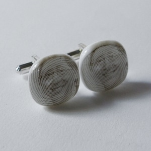 以下が含まれることがあります： 白とグレーのドット模様の正方形のカフスボタンのペア。カフスボタンは銀色の金属製の裏地が付いています。カフスボタンのデザインは、スパイラルまたは円形のパターンに見えます。