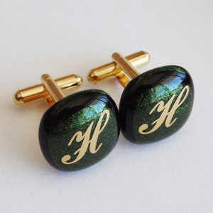 May include: A pair of gold cufflinks with black and green glass fronts. The glass is glittery and has a gold letter "H" engraved on each cufflink.