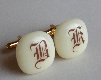 May include: A pair of white cufflinks with gold accents. The cufflinks feature a red, gothic-style letter "B" on one and a red, gothic-style letter "R" on the other.
