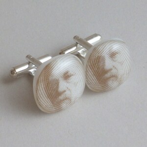 May include: A pair of silver cufflinks with white round faces featuring a brown patterned design of a face.