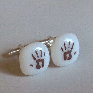 May include: A pair of white cufflinks with a brown handprint design. The cufflinks are square shaped and have a silver metal backing.