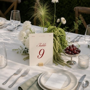 May include: A cream-coloured table number card labelled Table 9 with the names Adrian and Kaia and the date 25.06.2026 printed in red. The card is held upright by a small wooden block and is surrounded by white flowers and green foliage.