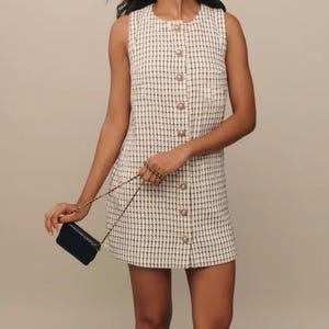 Peut inclure: Une robe sans manches à carreaux blancs et noirs avec un devant boutonné et une petite poche sur le côté gauche. La robe est associée à un sac à main noir à bandoulière chaîne et à des bottes noires mi-hautes.